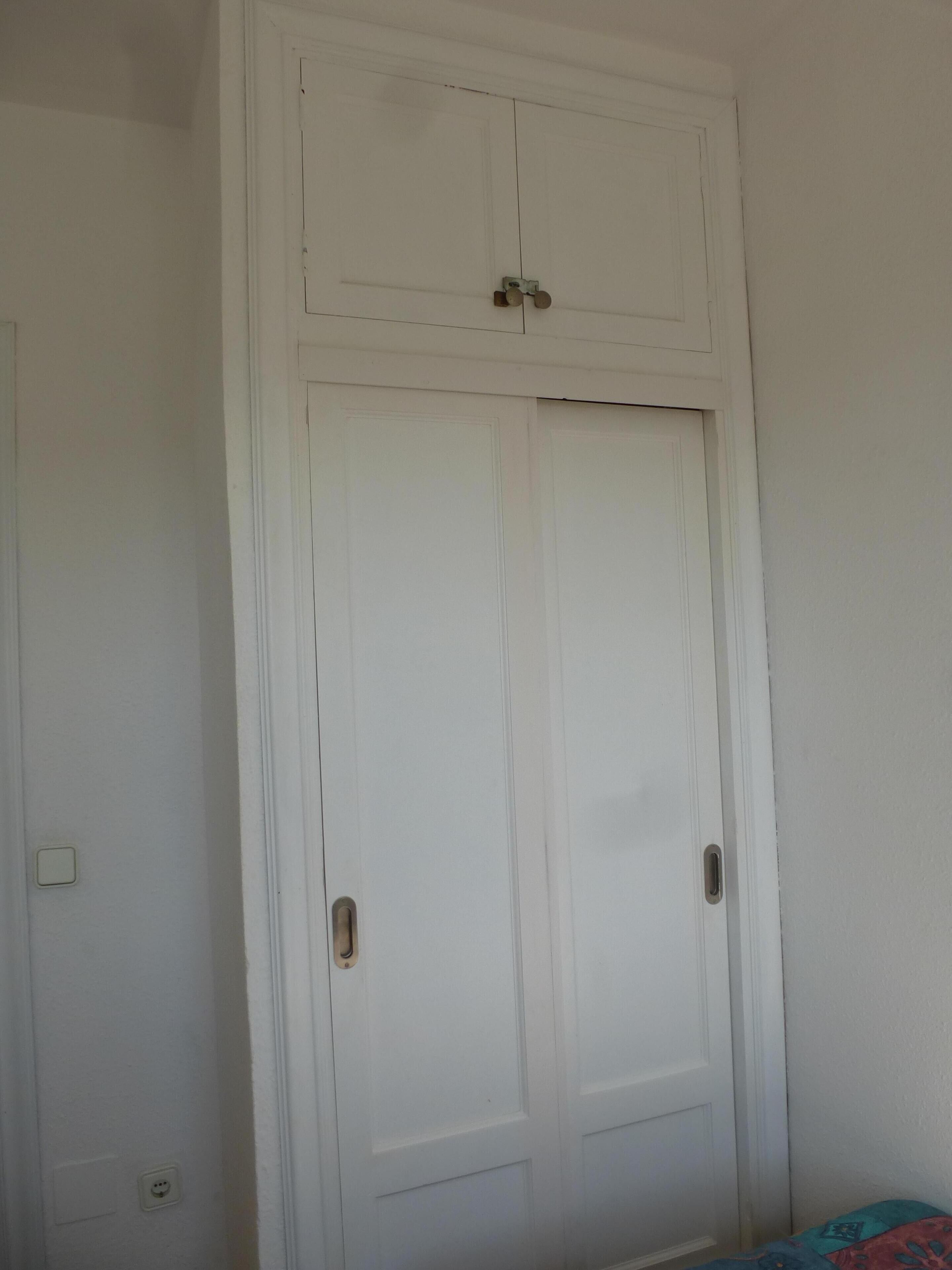 Une armoire blanche à deux portes, avec une petite étagère en haut.