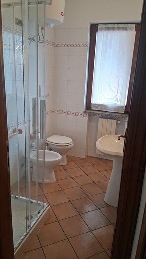 Une salle de bain avec une douche à parois de verre, deux toilettes, un lavabo et une fenêtre agrémentée de rideaux à motifs.
