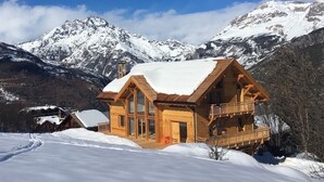 A wooden chalet with large windows and a balcony, surrounded by snow-covered mountains.
