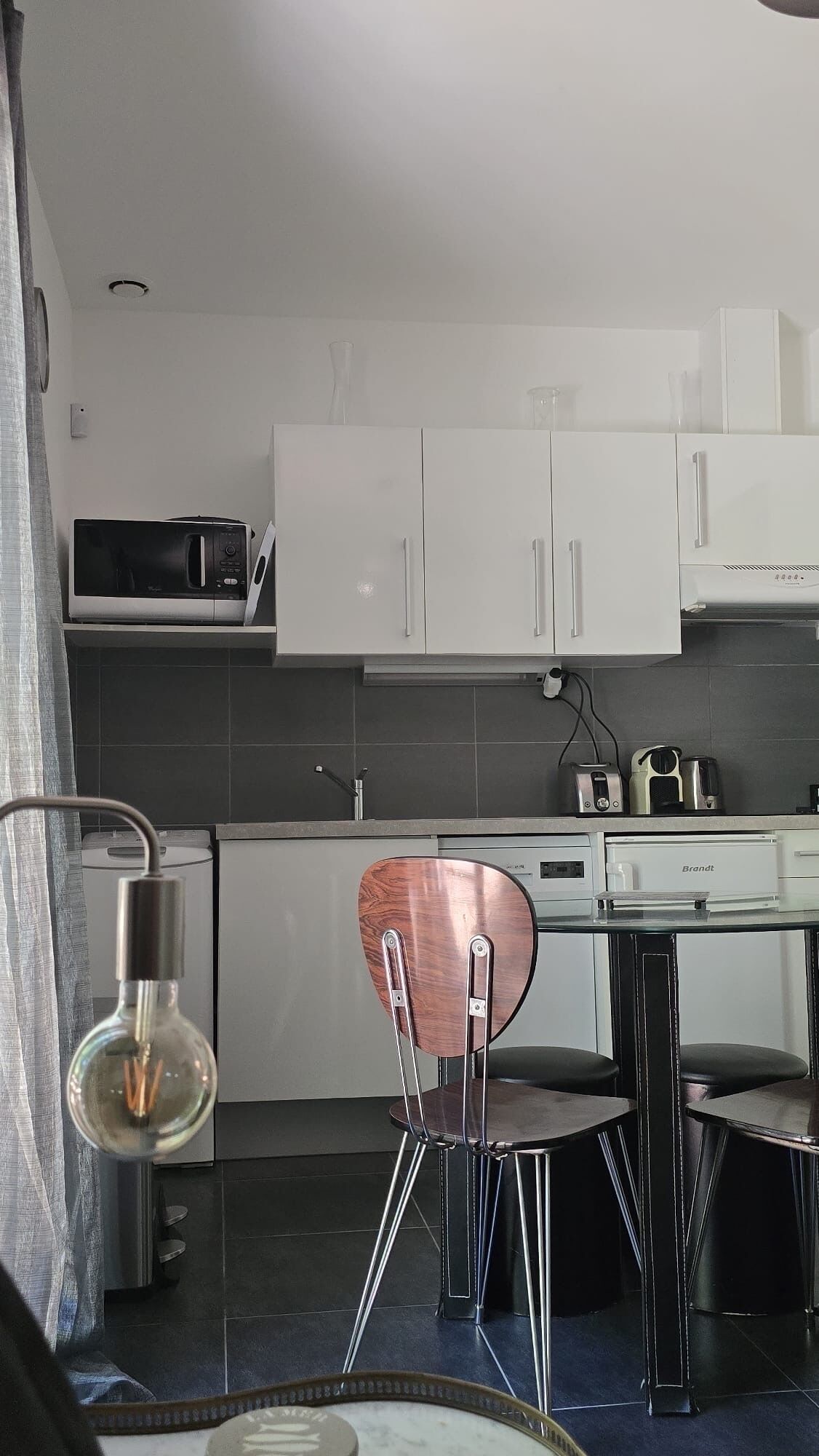 A compact kitchen with white cabinets, a microwave, a toaster, and a Brandt oven. There is a round wooden chair with metal legs and a small table.