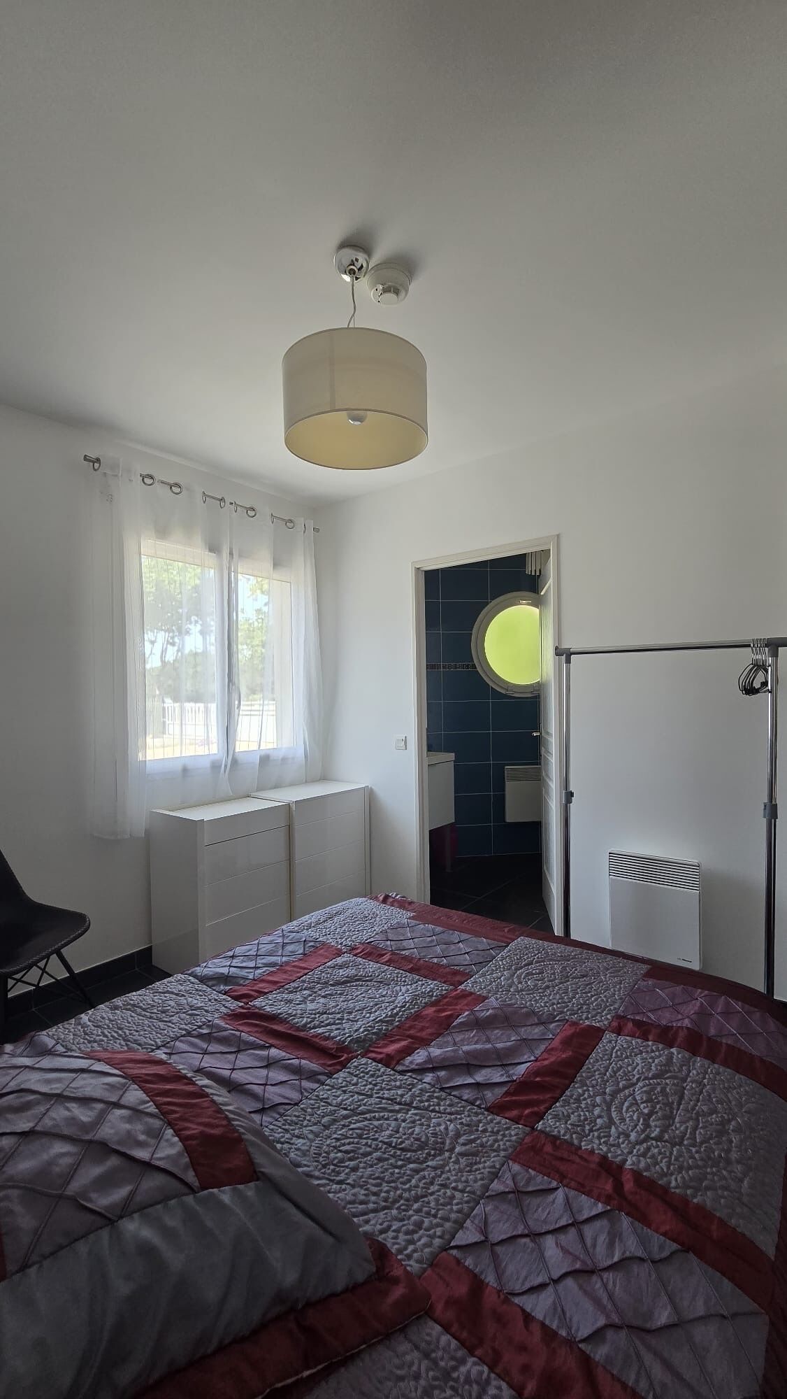 A bedroom with a bed, a chair, a window with curtains, a ceiling light, and a door leading to another room.