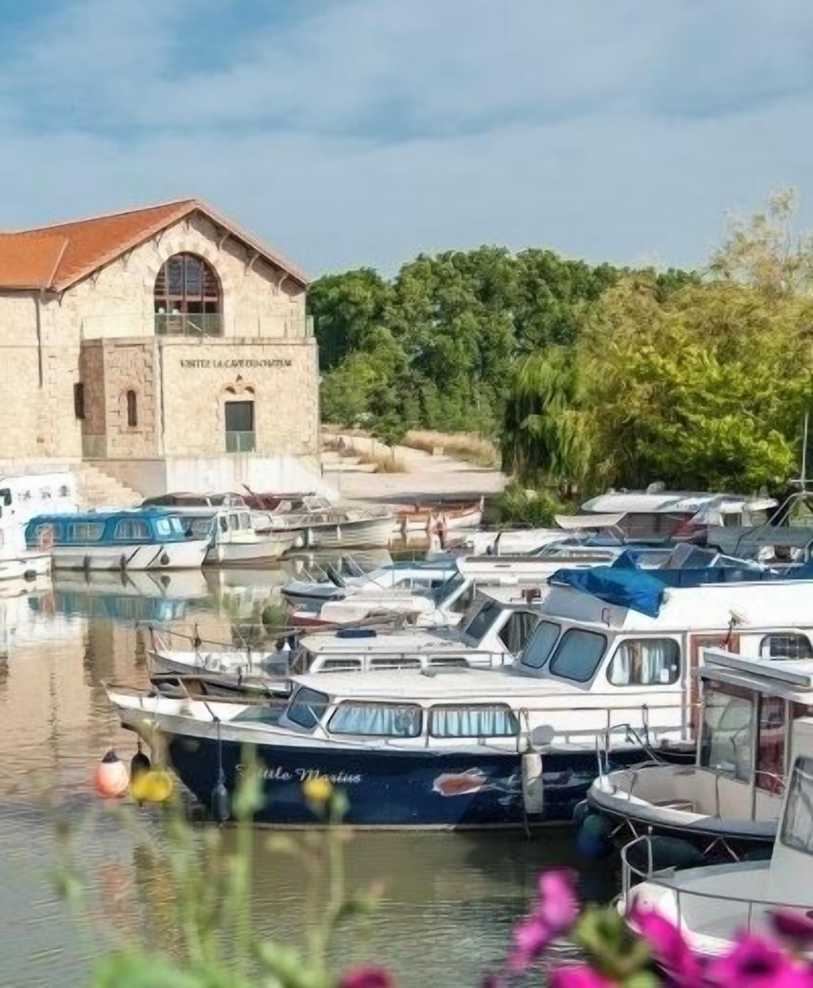 A marina with boats docked, a building with a stone facade, and lush greenery in the background.