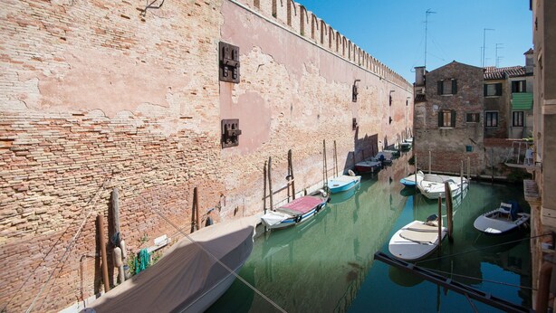Un canal étroit, bordé de bateaux amarrés, un mur de briques et un bâtiment surmonté d’un toit en tuiles.