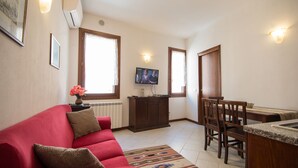 Un salon avec un canapé rouge, un téléviseur fixé au mur, un coin repas avec une table et des chaises en bois, et un coin cuisine avec un comptoir et des placards.