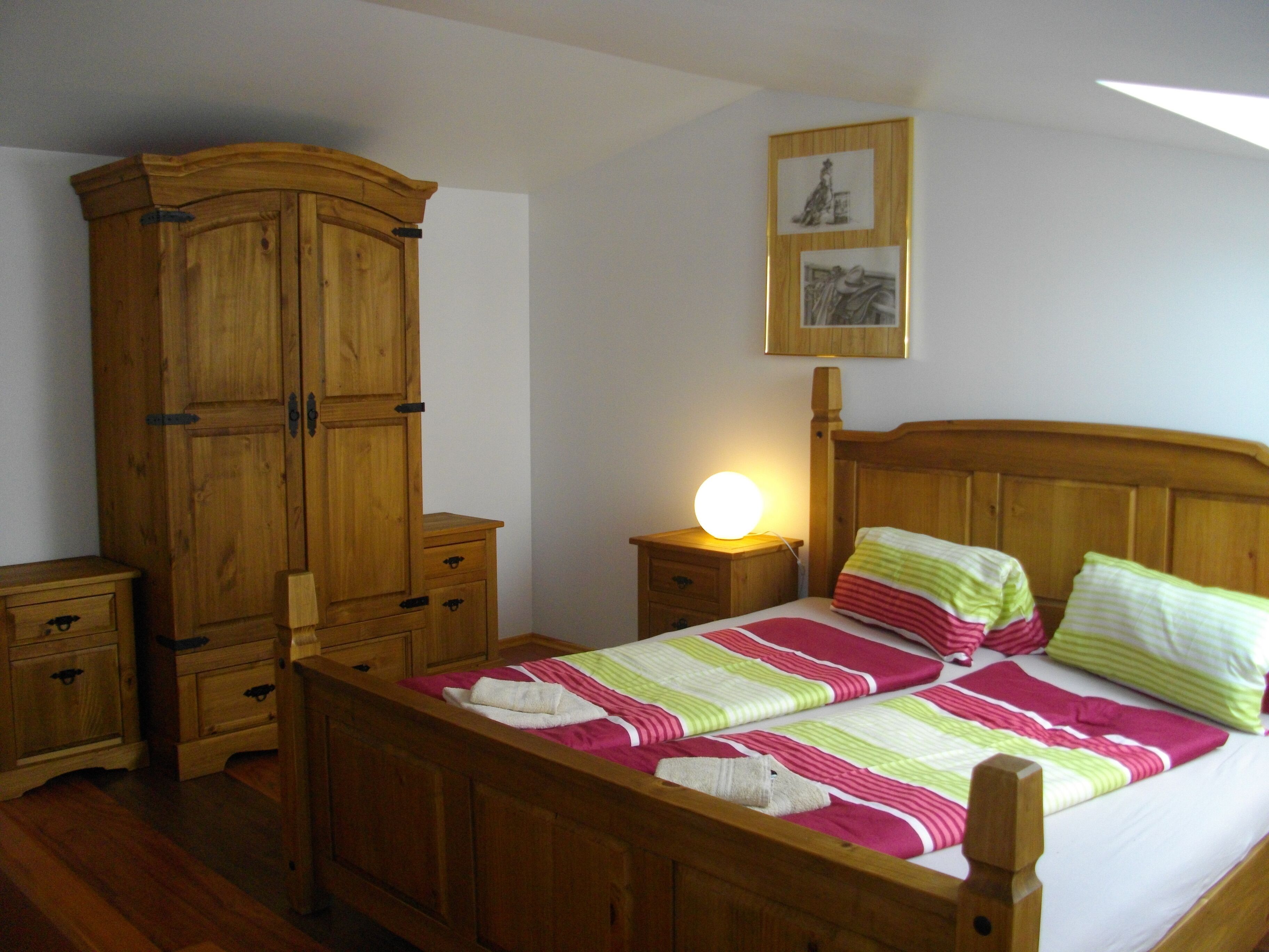 A bedroom with a wooden bed, a nightstand with a lamp, a wooden wardrobe, and a framed picture on the wall.