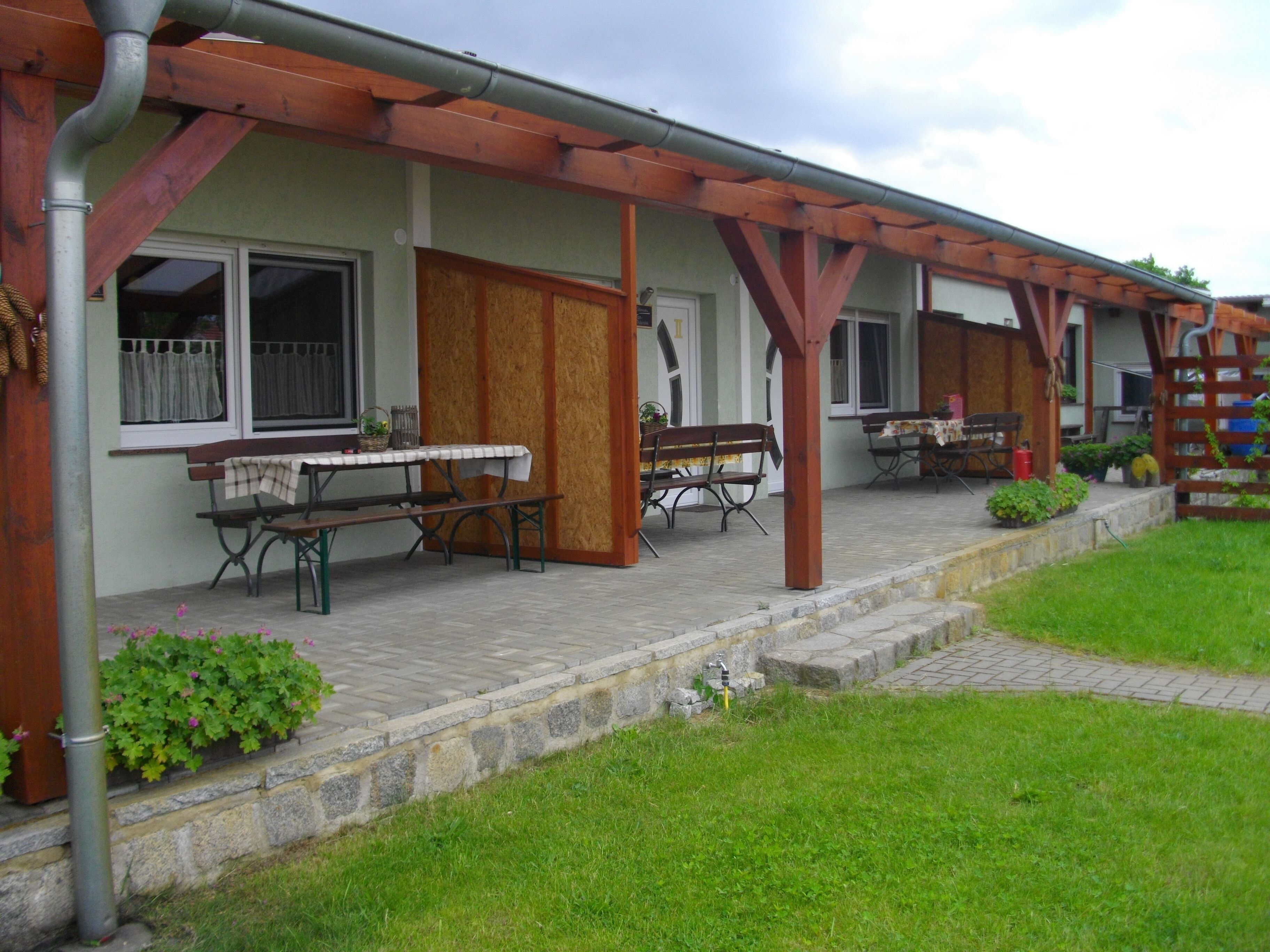 A covered outdoor area with a picnic table and benches, stone steps leading to a building, and a grassy lawn.