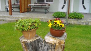 A building with a wooden porch, a table, and chairs. There are two flower pots on a tree stump. - VierJahreszeiten Appartments (Lübben (Spreewald))
