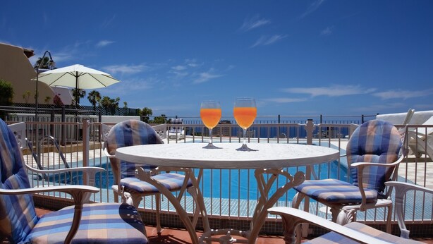 Een zwembadgedeelte met ligbedden en een tafel, twee glazen oranje drank, en een heldere blauwe lucht.