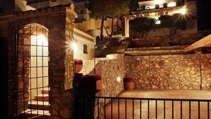 Ein Innenhof mit Steinwänden, einem Metallgitter, Blumentöpfen und einer Treppe, die zu einem Gebäude mit beleuchteten Fenstern führt.