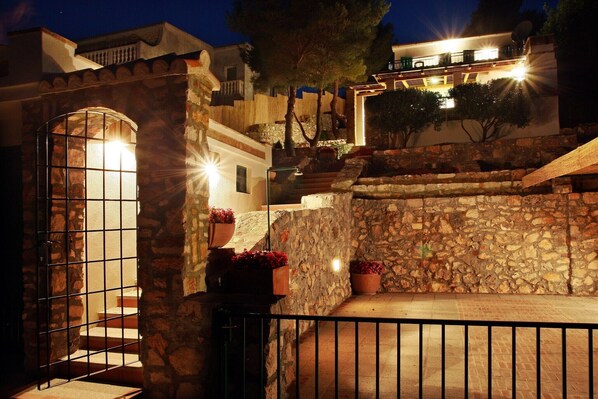 A stone-walled courtyard with a metal gate, potted plants, and a staircase leading to a building with illuminated windows.