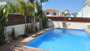 A swimming pool with a wooden deck, surrounded by tropical plants and a white wall with a wooden fence. -   Sea  and Sun rise views. Villa with private pool and gated children Play Area. (paralimni)