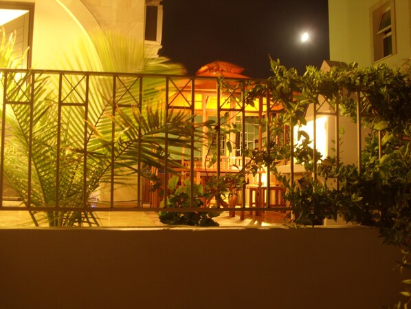 A nighttime view of a building with a balcony, surrounded by greenery and a fence.