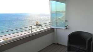 A balcony with a view of the sea, a chair, and a window with a view of the beach and ocean.