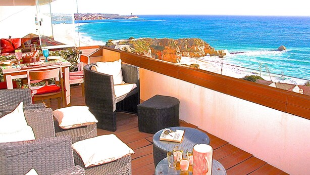 A balcony with wicker furniture, a table with drinks, and a view of the sea and cliffs.