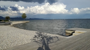 Un muelle de madera que conduce a una playa de guijarros, con vista al mar y montañas distantes bajo un cielo nublado.
