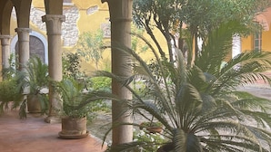 Un patio cubierto con columnas de piedra, plantas en macetas y un edificio amarillo al fondo.