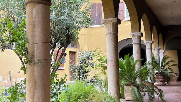 Un patio cubierto con columnas de piedra, plantas en macetas y suelo de baldosas.