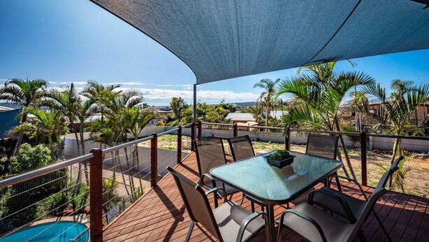 Un patio techado al aire libre con mesa y sillas, con vista a una piscina y palmeras.