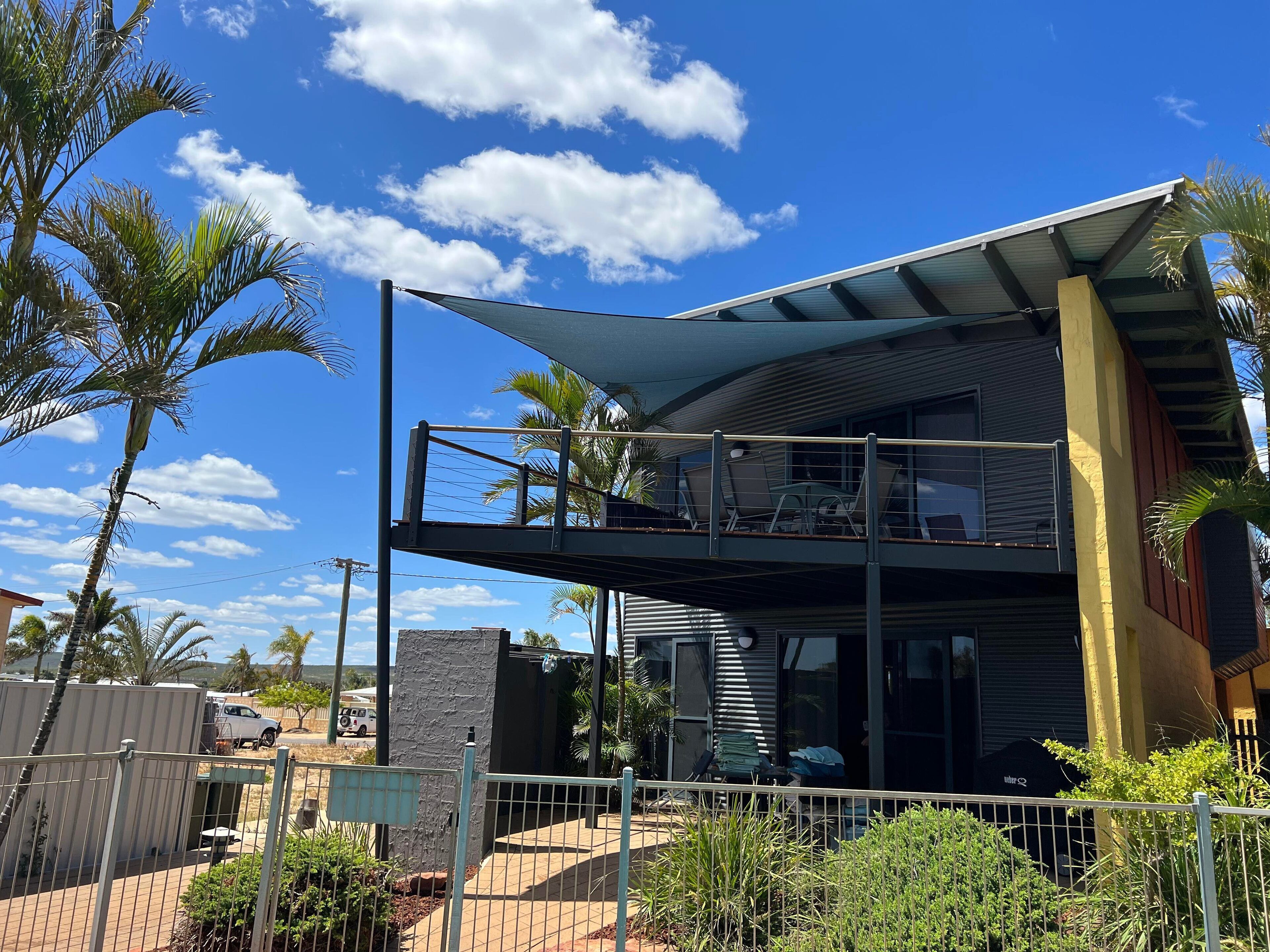 A modern two-story building with a balcony, surrounded by palm trees and a clear blue sky.
