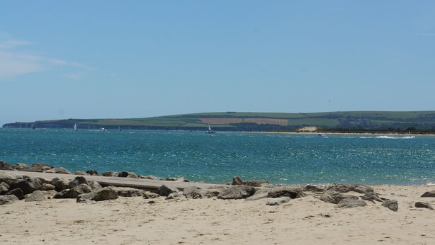 A coastal beach with a rocky shoreline, a clear blue sea, and distant hills. - Your holiday is just 100m from award winning beaches on Sandbanks peninsular. (Poole)