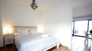 Une chambre à coucher comprenant un lit, des tables de chevet, une commode, une lampe suspendue et une vue sur l’extérieur par une porte.