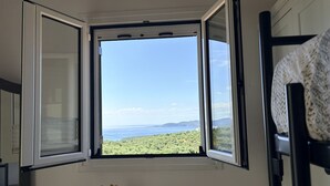 Ein Zimmer mit Fenster und Blick auf eine Küstenlandschaft, ein kleiner Tisch mit Blumenvase sowie ein Etagenbett.