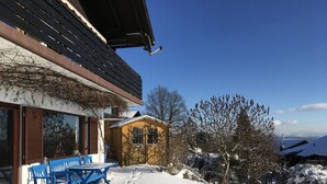 A wooden building with a sloped roof, a small porch with blue chairs, and a snow-covered ground. - Adventure and Relaxation for you and your family in the Southern Bavarian Forest (Windorf)