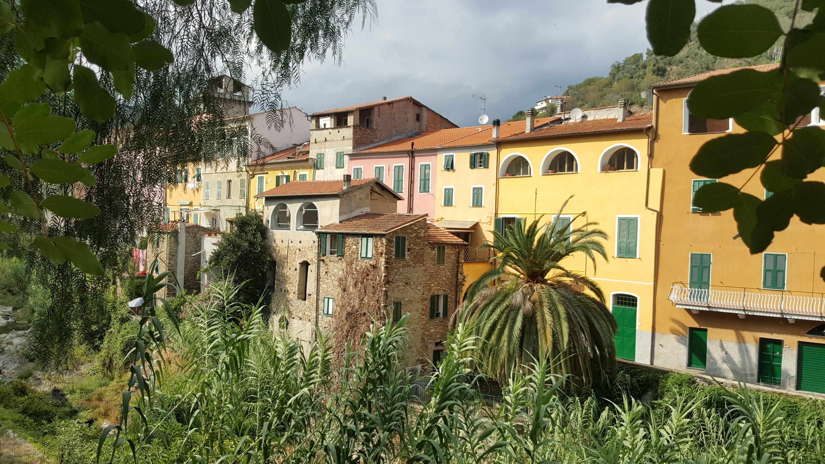 A row of colorful buildings with green shutters, a stone building with ivy, and lush greenery in the foreground.