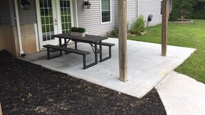 Une terrasse couverte avec une table de pique-nique et des bancs, une porte blanche avec des panneaux de verre vert et une petite plante en pot.
