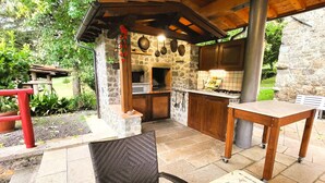 An outdoor kitchen area with stone walls, a wooden table, and a wicker chair. - 4 bedroom, 2 bathroom converted Mill house with private pool.  Outdoor kitchen! (Poggio Camporgiano)