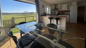 Une cuisine moderne dotée d’une grande fenêtre, un coin repas avec une table en verre et une vue sur un champ verdoyant.