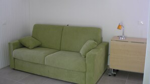 Un canapé vert avec deux coussins, une petite table d’appoint en bois avec une lampe, et un sol carrelé.