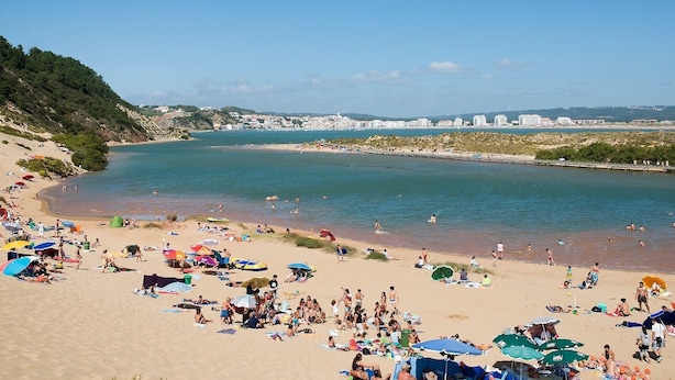 Een strand met mensen die zonnebaden en zwemmen, een pier en gebouwen op de achtergrond.