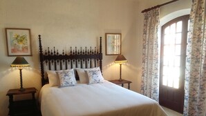 Een net opgemaakt bed met bloempatroon kussens, twee nachtkastjes met lampen, en een groot raam met bloemen gordijnen.