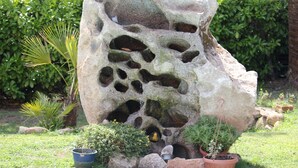 Une grande formation rocheuse percée de nombreux trous, entourée de plantes en pot et de verdure.