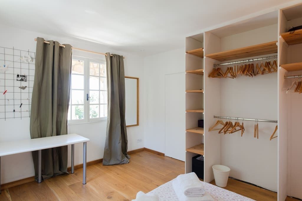 A room with a wooden floor, a white desk, a window with curtains, and a walk-in closet with wooden shelves and hangers.
