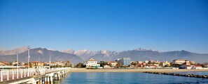 Un pueblo costero con un muelle que se adentra en el mar, edificios con techos rojos y montañas al fondo.