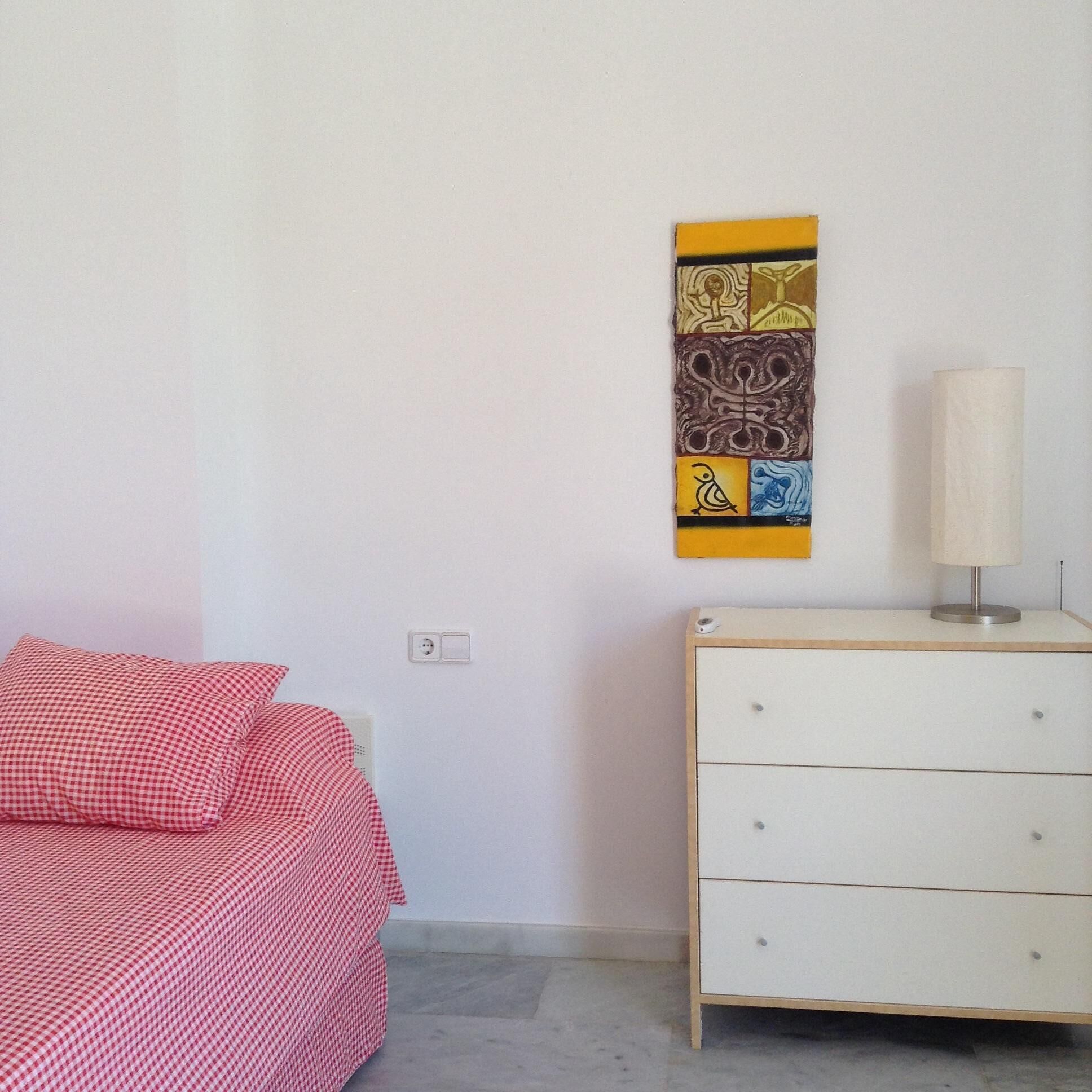 Une chambre avec un lit à motifs à carreaux, une commode blanche et une œuvre d’art abstraite accrochée au mur.
