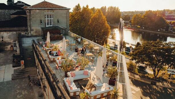 En udendørs terrasse med siddepladser og borde med udsigt over en flod, beliggende ved siden af en historisk bygning.