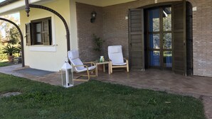 Een overdekt terras met twee witte lounge fauteuils, een bijzettafeltje en een potplant.