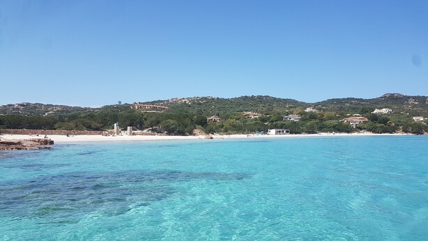 A coastal view with clear turquoise waters, a sandy beach, and buildings on a hillside in the background. - Maison "The Little Gate", seaside paradise. (porto san paolo)