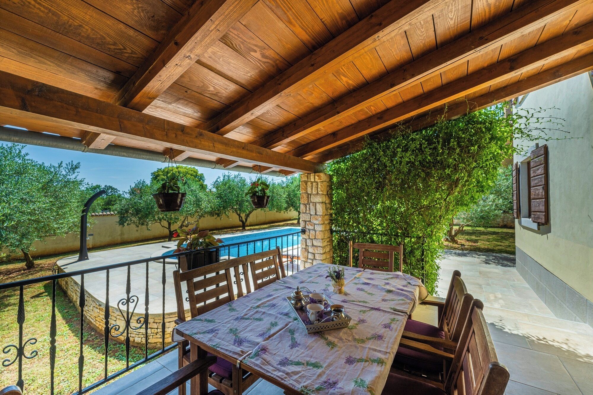 Un patio al aire libre con una mesa de madera dispuesta para comer, rodeada de sillas, con vistas a una piscina y abundante vegetación.