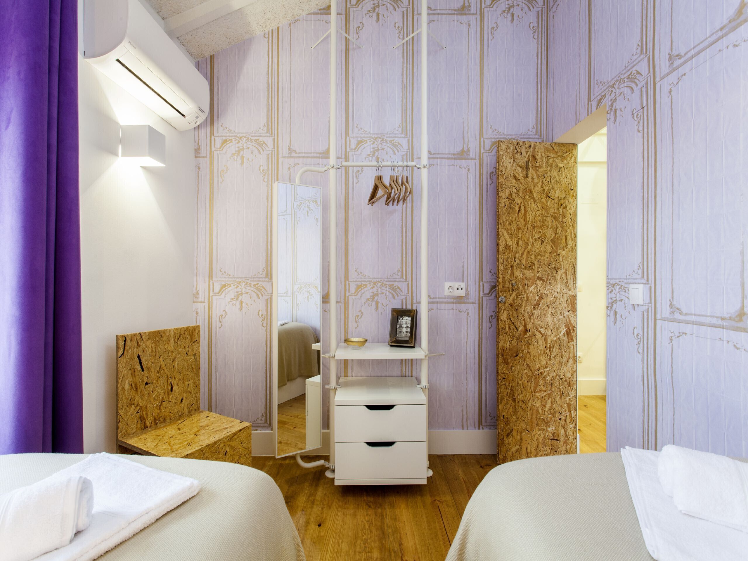 A modern bedroom with two beds, a wooden bench, a vanity with a mirror, and a wall-mounted shelf with a picture frame.