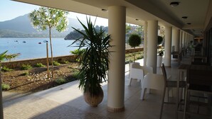 Um espaço ao ar livre coberto com vista para o mar, plantas em vasos e cadeiras brancas.