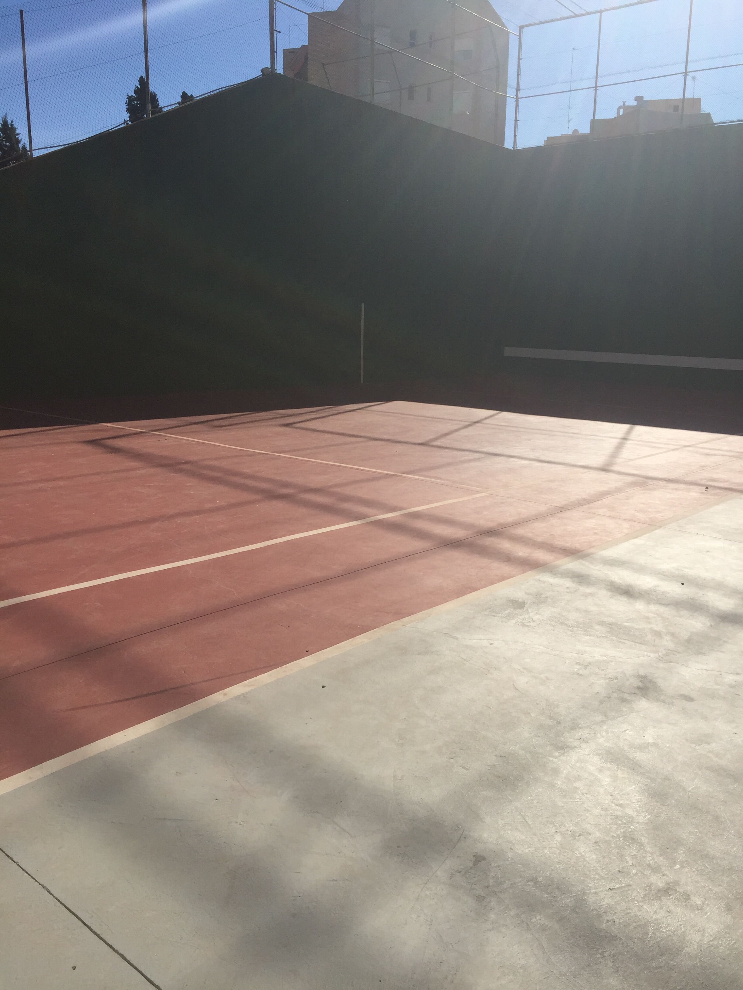 A tennis court with a net, surrounded by a fence and buildings in the background.