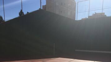 A tennis court with a net, surrounded by a fence and buildings in the background.