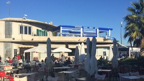 Un café al aire libre con mesas y sillas, un edificio con un letrero azul al fondo, y una palmera a la derecha.