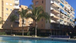 Una piscina con bordes azulejados, rodeada de una zona pavimentada. En el fondo, edificios de varios pisos con balcones y un cielo azul despejado.