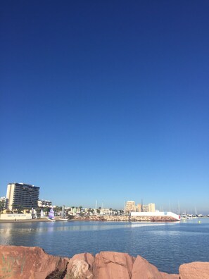 Un paisaje urbano costero con cielo despejado, un puerto deportivo con barcos y edificios a lo largo de la costa.