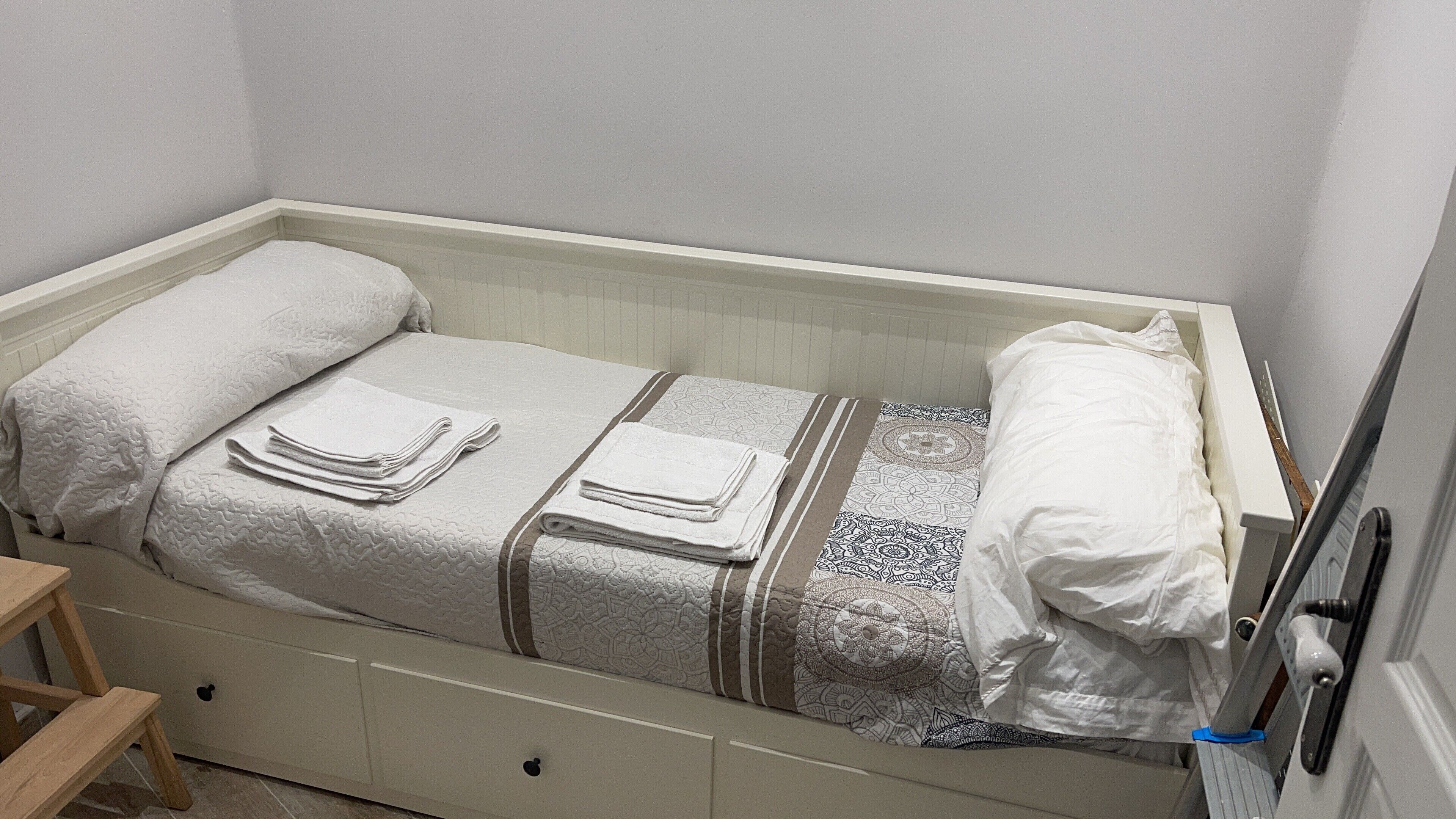 Una cama individual con colcha blanca, dos almohadas y toallas dobladas.